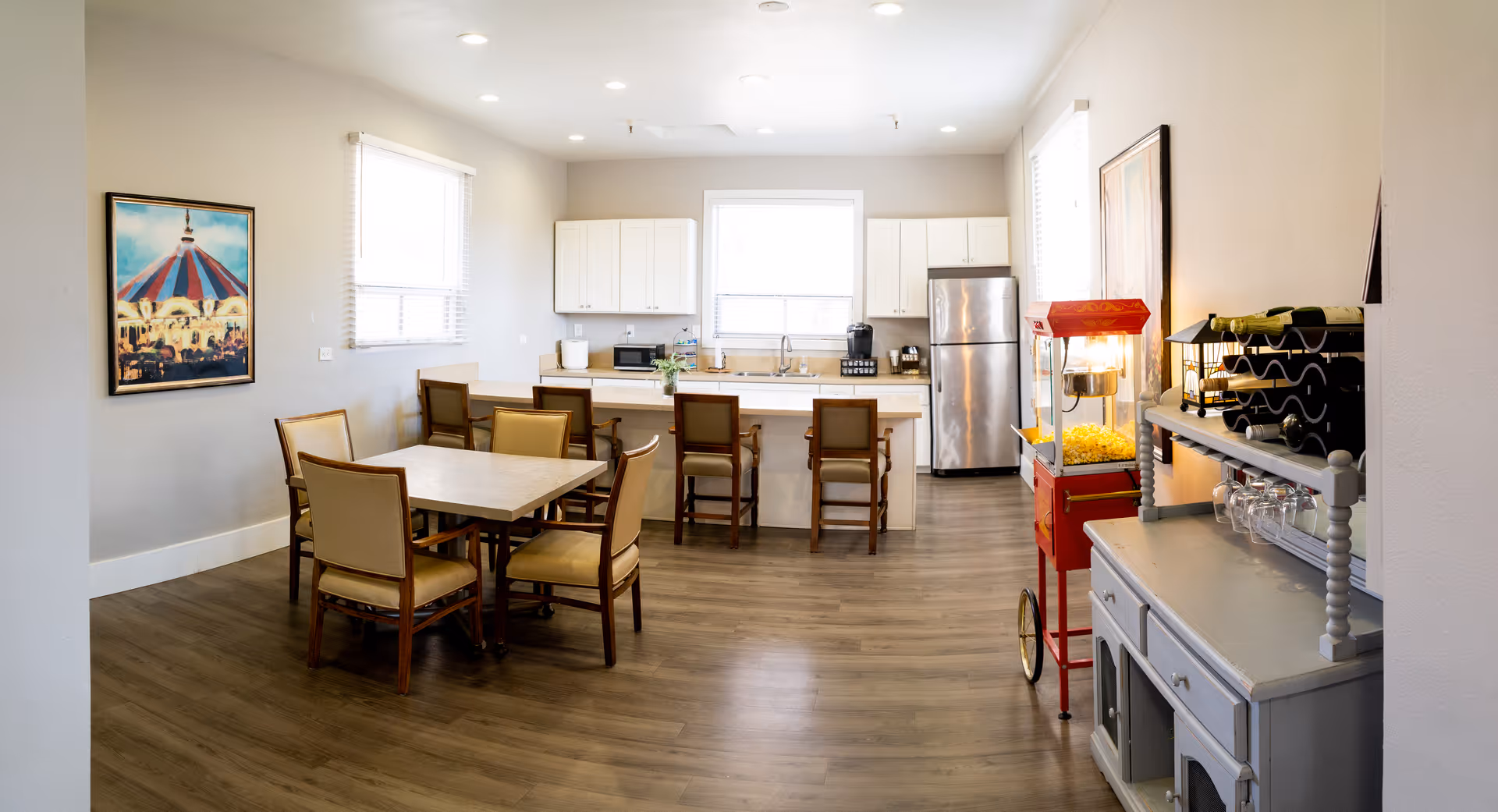 A bright and clean kitchen and dining area in a senior living facility. The room features a dining table with six chairs, a kitchen island with four bar stools, white cabinets, a stainless steel refrigerator, a popcorn machine, and a wine rack with glasses. There are two windows with blinds allowing natural light to fill the space, and framed artwork on the walls.