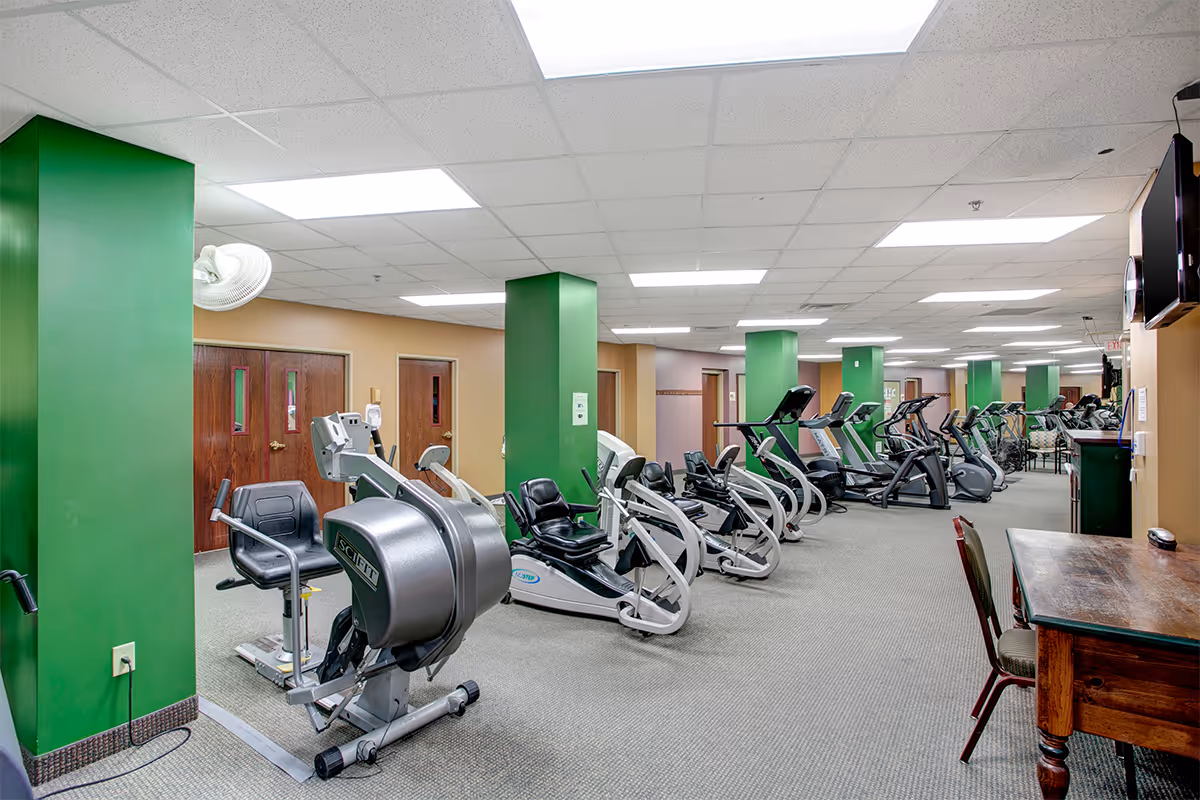 Indoor fitness room with multiple exercise machines including recumbent bikes and elliptical trainers. The room has green pillars, beige walls, a carpeted floor, and fluorescent ceiling lights. There is a wooden table with chairs on the right side and a wall-mounted TV.