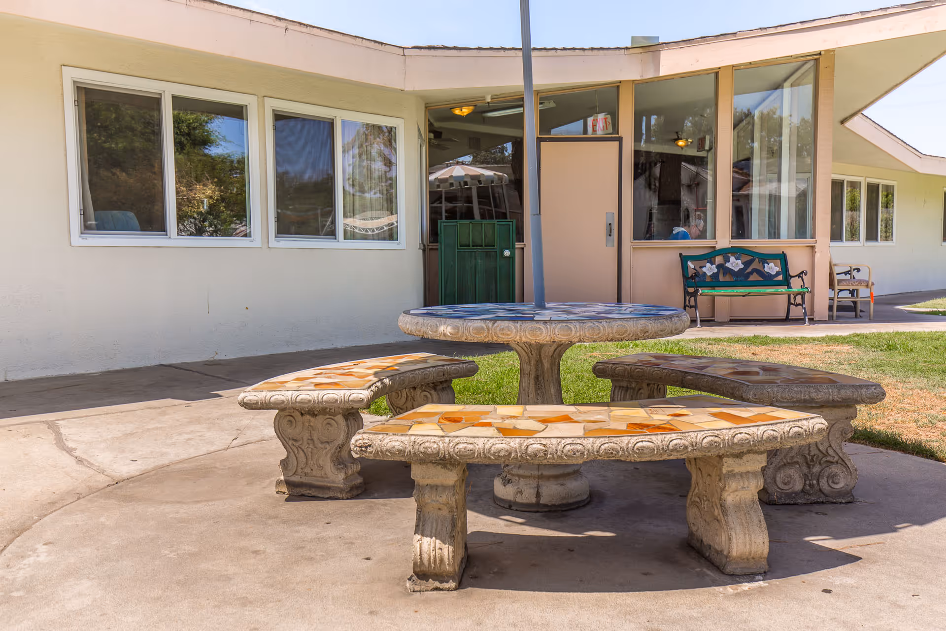 Outdoor seating area with a round stone table and three curved stone benches on a concrete patio. The background shows a building with multiple windows, a door, and a bench with a decorative backrest. There is grass adjacent to the patio area.