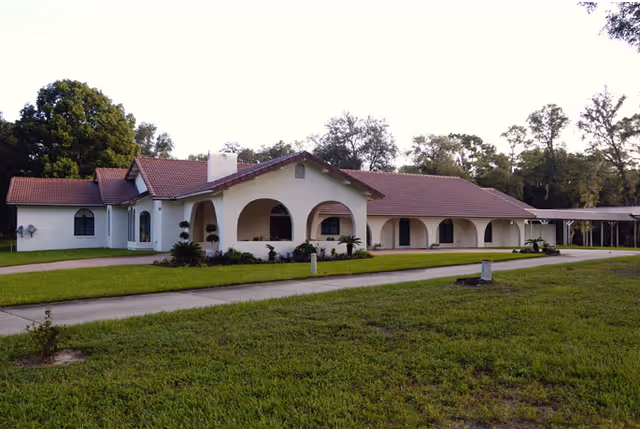 Single-story building with white walls and a red tiled roof surrounded by green grass and trees, featuring arched windows and a covered walkway on the right side.