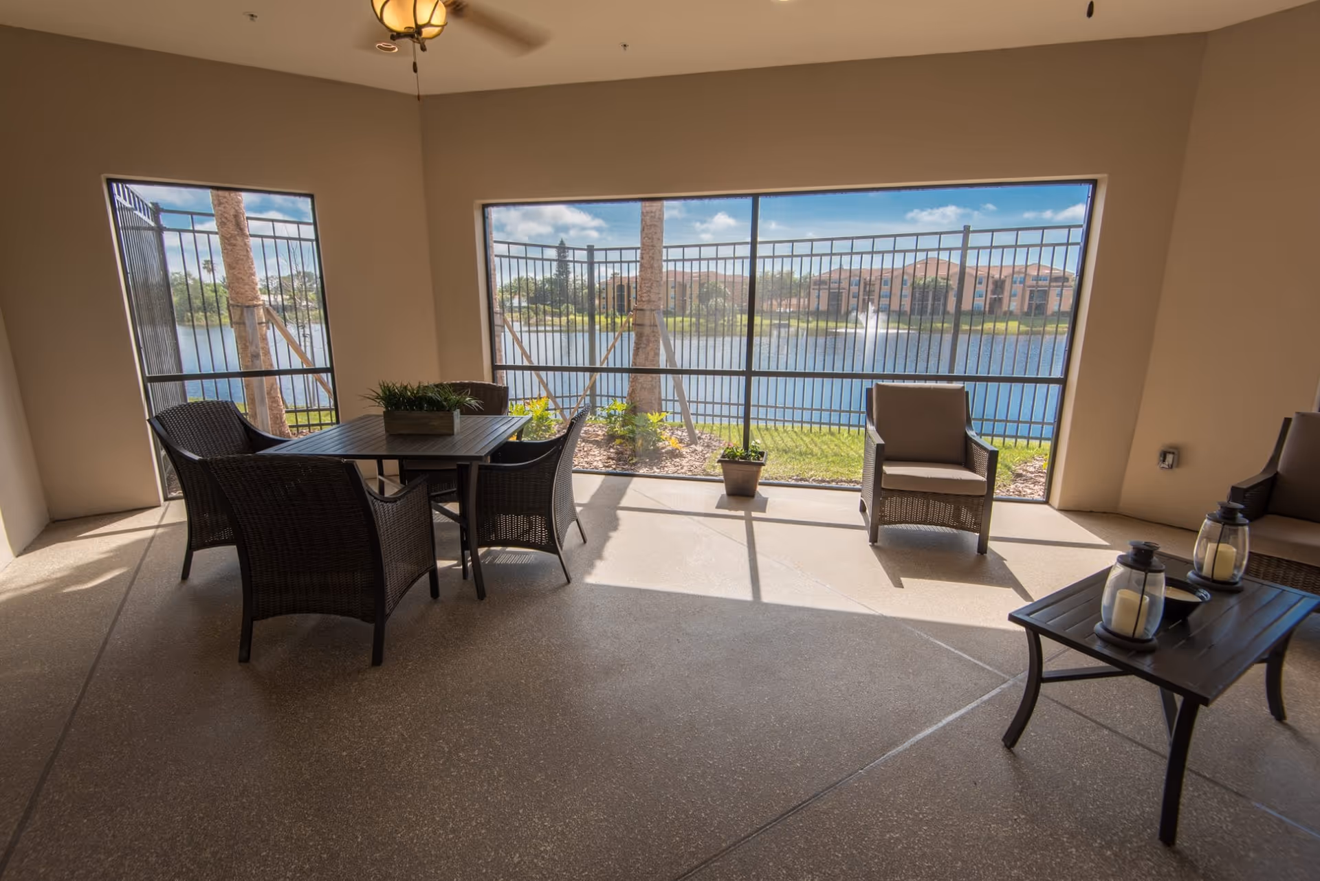 A covered patio area with a table and four wicker chairs, two additional wicker armchairs, and a small coffee table with two candle lanterns. The patio overlooks a fenced lake with a fountain and buildings in the background under a blue sky with some clouds.