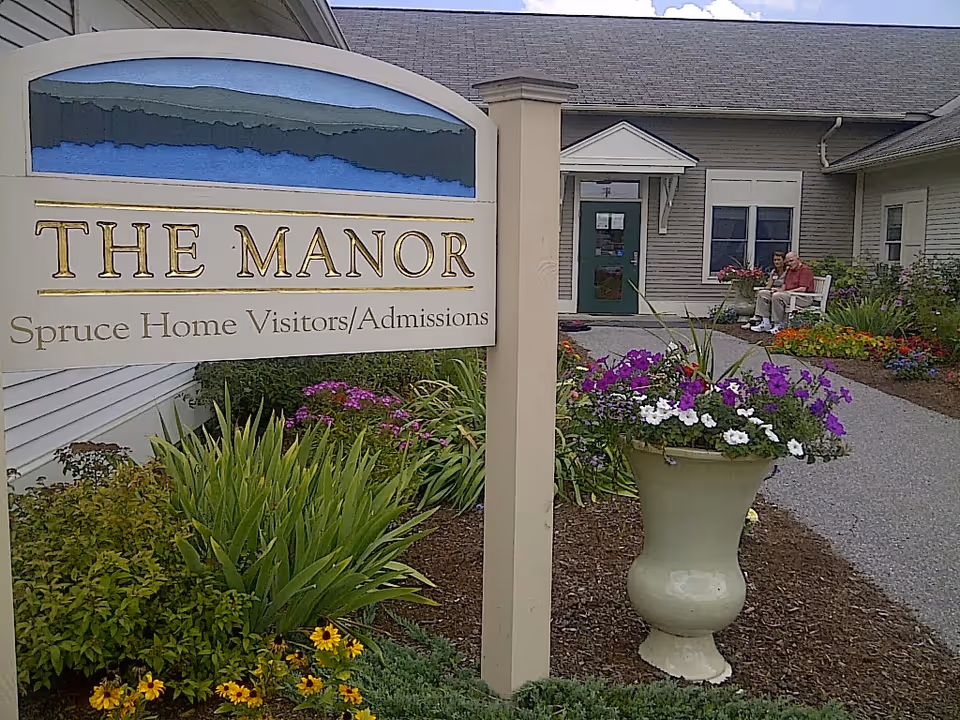 Front entrance of The Manor with a prominent sign, colorful flower planters, and two people seated on a bench near the doorway.