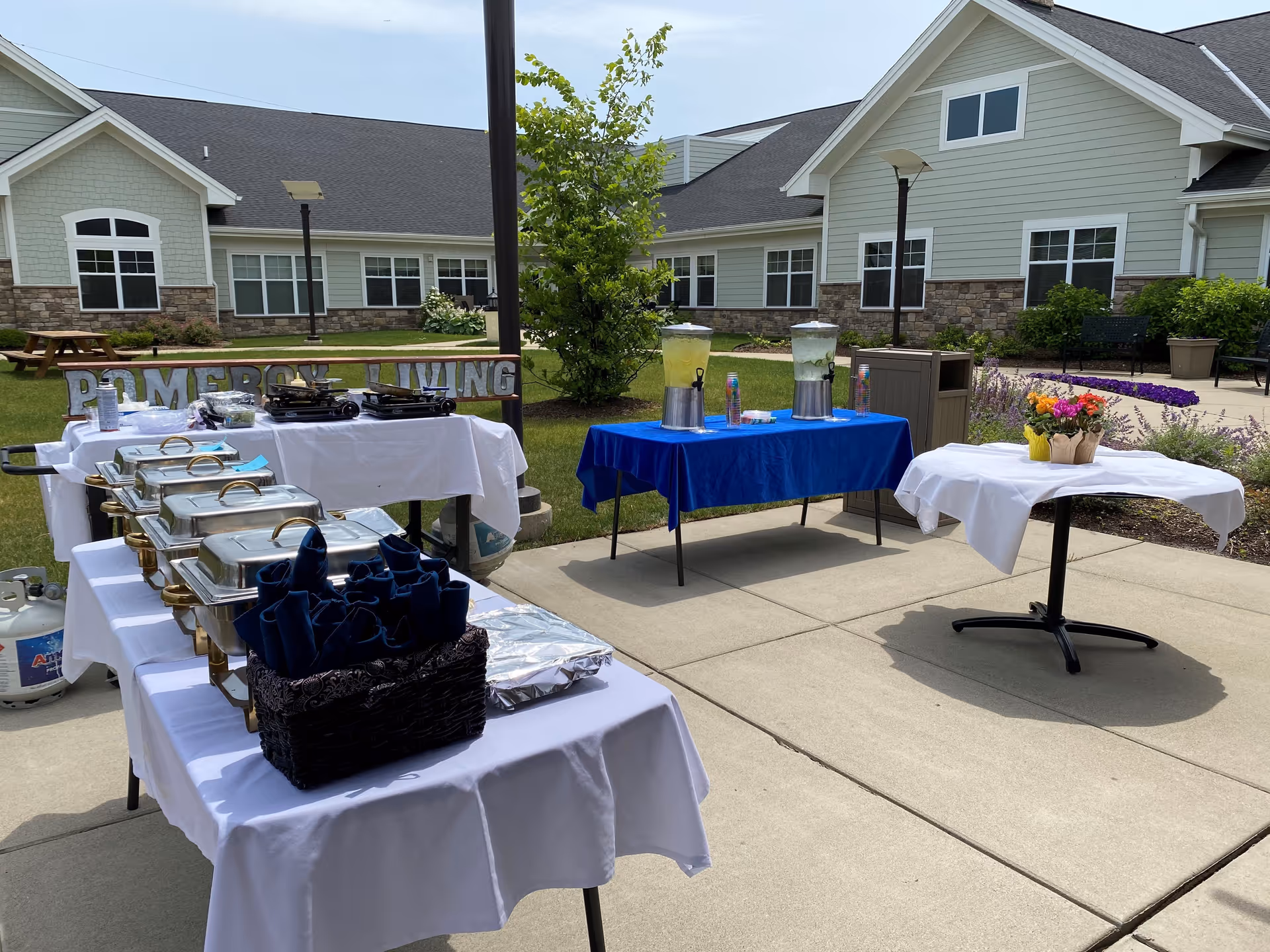 Outdoor courtyard set up with buffet tables, chafing dishes, drink dispensers, and flowers in front of a senior living building.