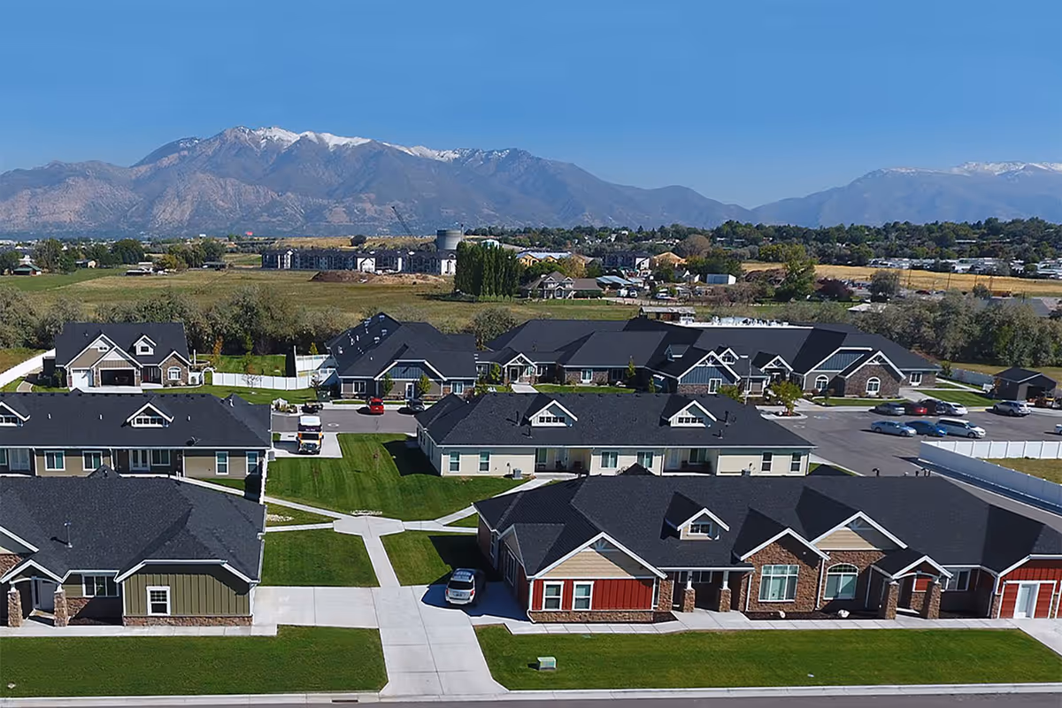 Aerial view of Cogir Of Lotus Park Senior Living facility showing multiple single-story residential buildings with dark roofs, green lawns, driveways, and parked cars. In the background, there are open fields, other buildings, and a mountain range with some snow on the peaks under a clear blue sky.