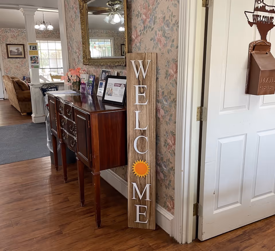 Interior view of a senior living facility with a wooden sideboard against a floral wallpapered wall. On the floor leaning against the wall is a wooden sign with the word 'WELCOME' written vertically, featuring a sun symbol in place of the letter 'O'. A large mirror hangs above the sideboard, reflecting a ceiling fan and window. To the right, a white door with a copper mailbox and a Texas-shaped welcome sign is partially open. In the background, a living room area with a brown couch and large windows is visible.