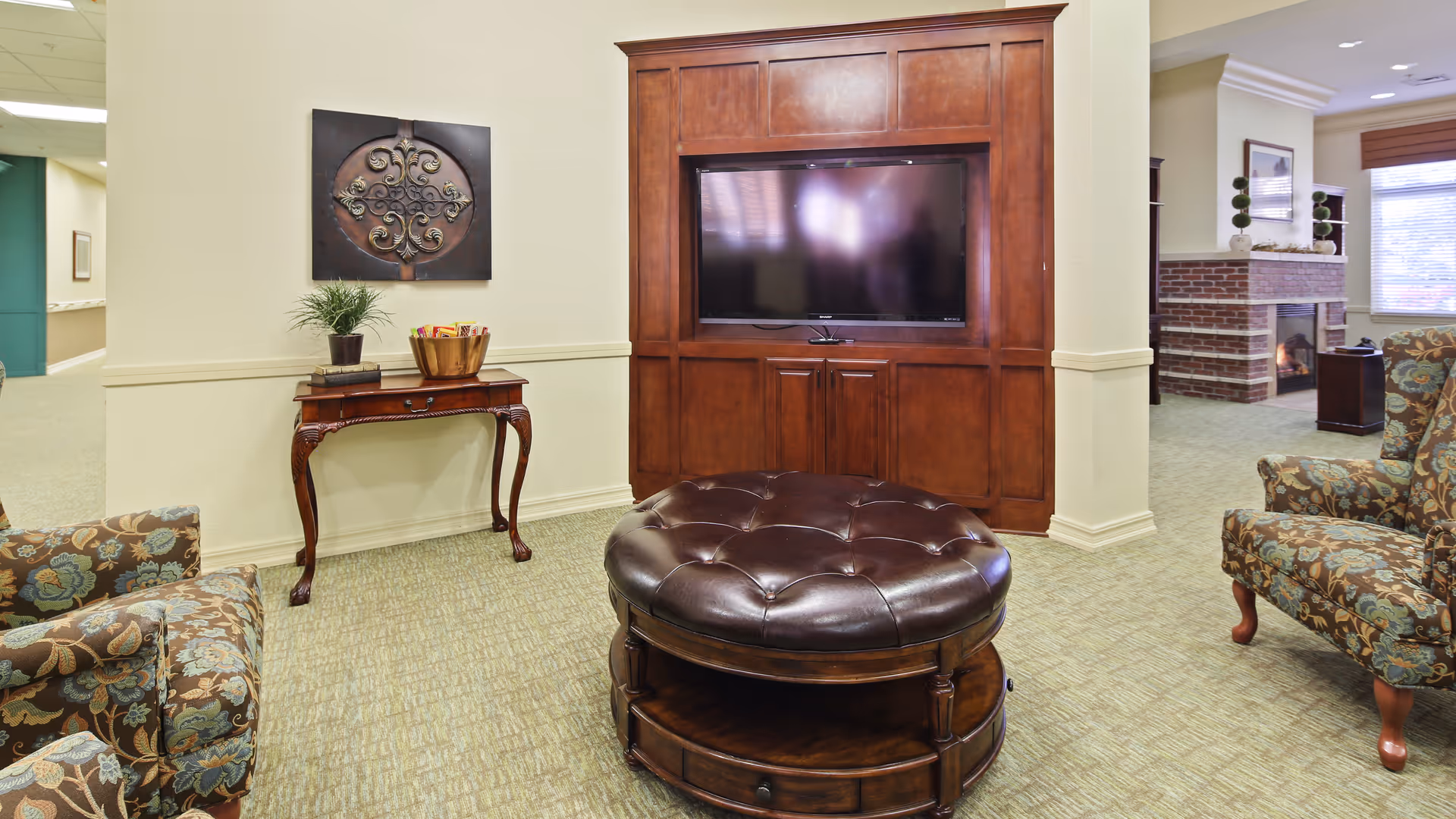 A cozy living room area in an assisted living facility featuring a large wooden entertainment center with a flat-screen TV, two floral upholstered armchairs, a round tufted leather ottoman with storage, a small wooden table with a plant and decorative bowl, and a brick fireplace in the background.