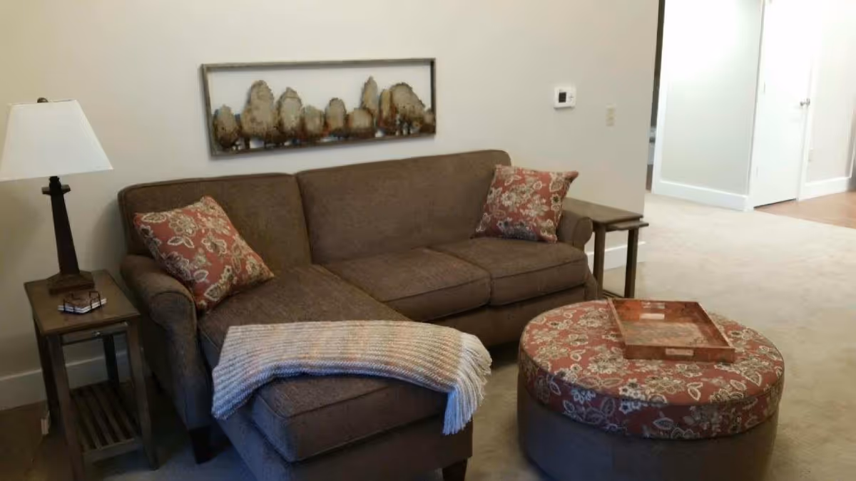 A cozy living room area with a brown sectional sofa adorned with two floral patterned pillows and a light-colored throw blanket. There is a round ottoman with a matching floral pattern and a wooden tray on top. A wooden side table with a lamp and a pair of glasses sits next to the sofa. The walls are light-colored with a decorative metal wall art piece above the sofa. A thermostat is visible on the wall, and a hallway with doors is seen in the background.