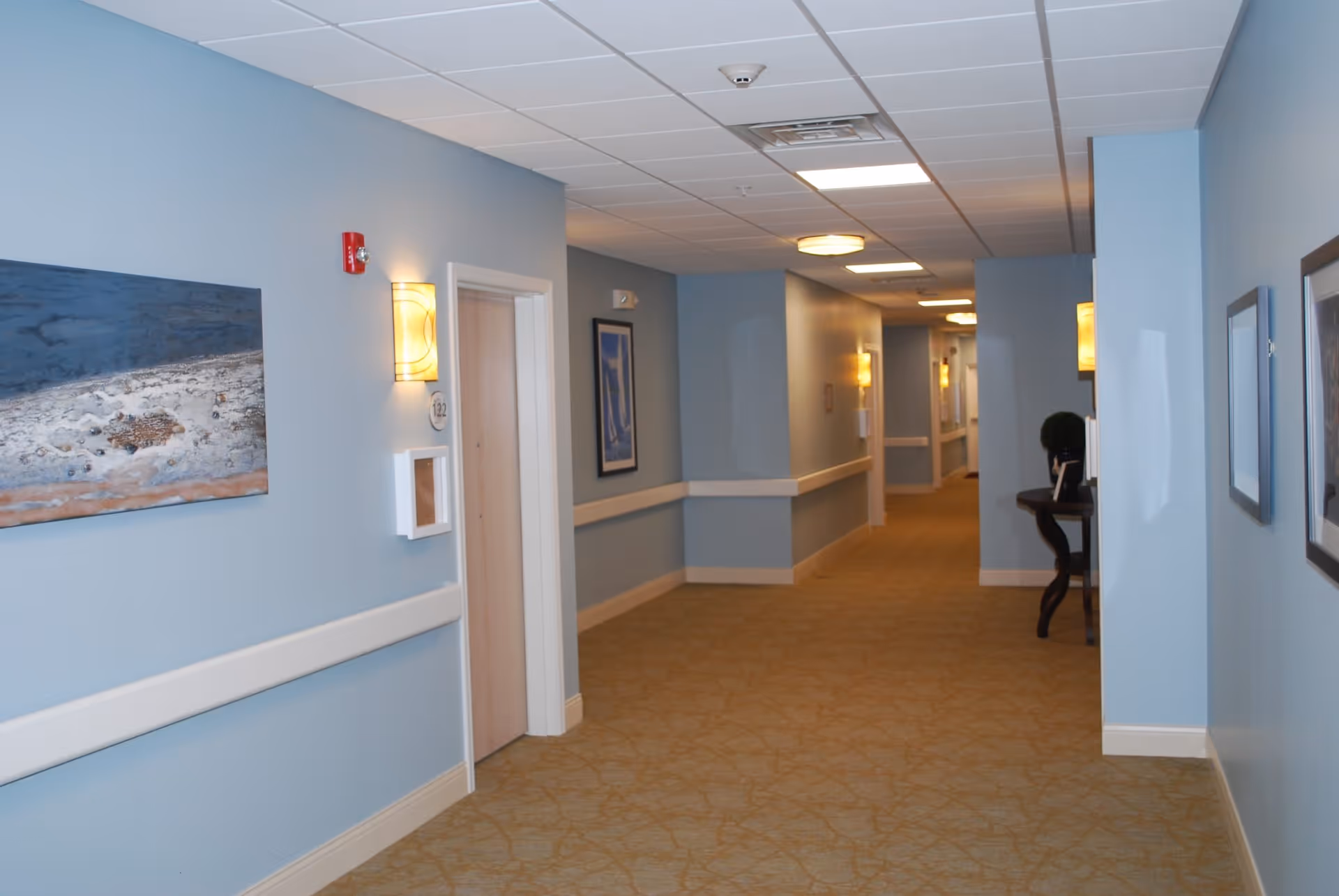 A well-lit hallway in a senior living facility with light blue walls, beige carpet, and several closed doors along the corridor. The walls are decorated with framed artwork and wall-mounted lights. A small table with a plant is visible on the right side of the hallway.
