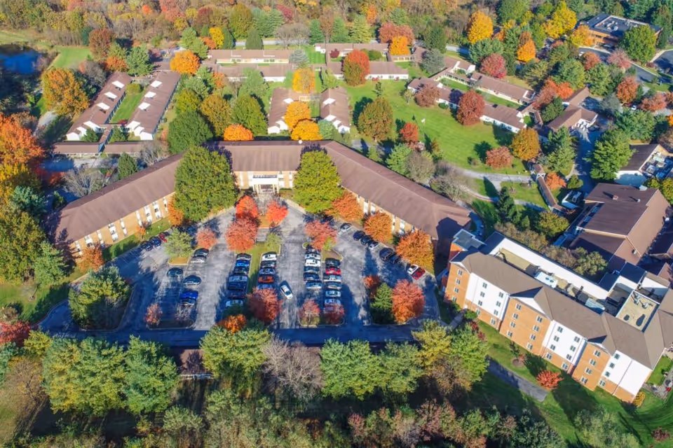 Aerial view of Heart Lands Senior Living Village At Ellicott City showing multiple buildings surrounded by trees with autumn foliage, a parking lot with cars, and green lawns.