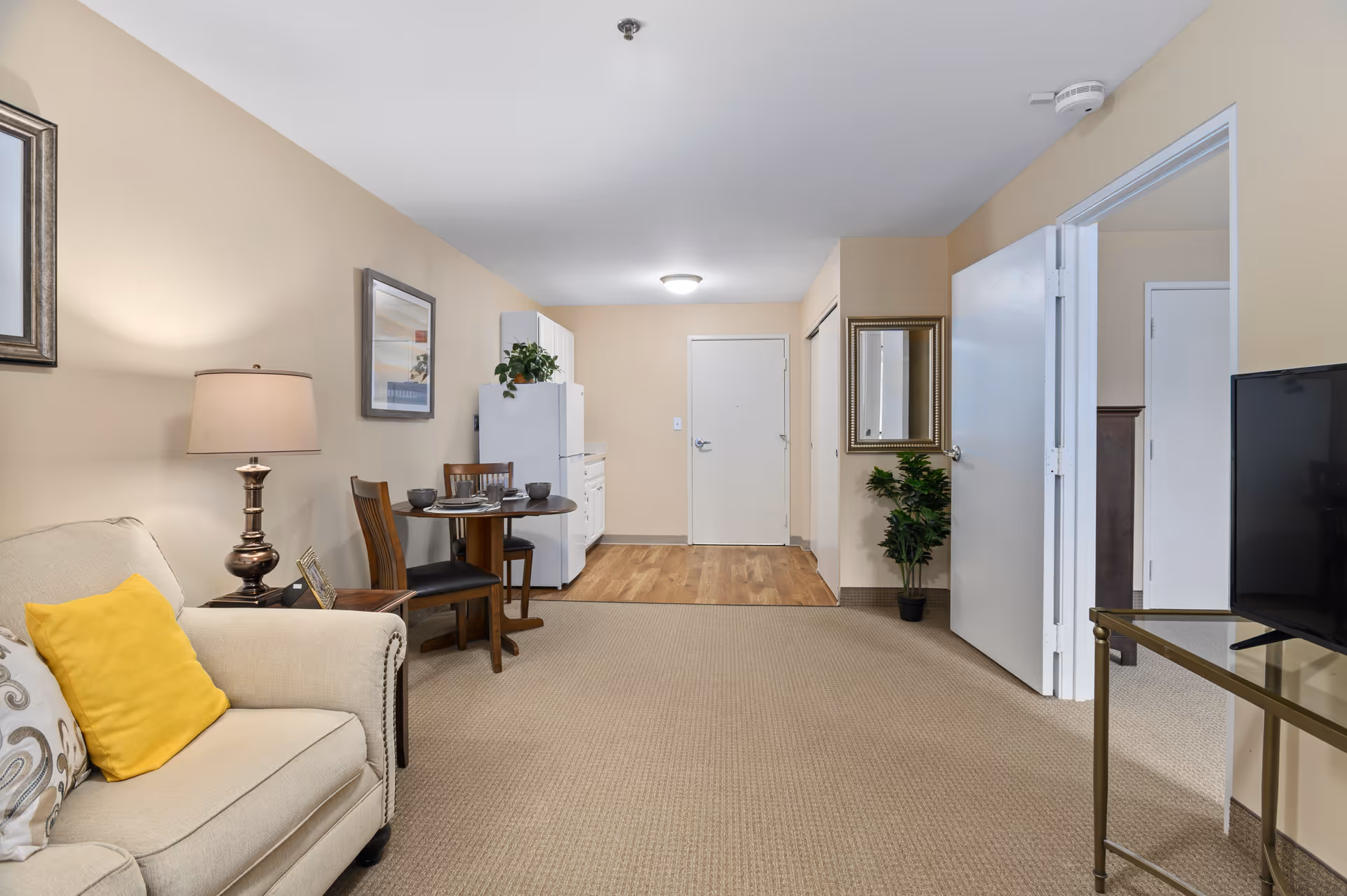 Interior view of a senior living apartment showing a beige sofa with yellow and patterned pillows, a wooden side table with a lamp, a small round dining table with two chairs, a white refrigerator and cabinets in the background, a hallway with a closed white door, a wall mirror, a potted plant, and a TV on a glass stand.