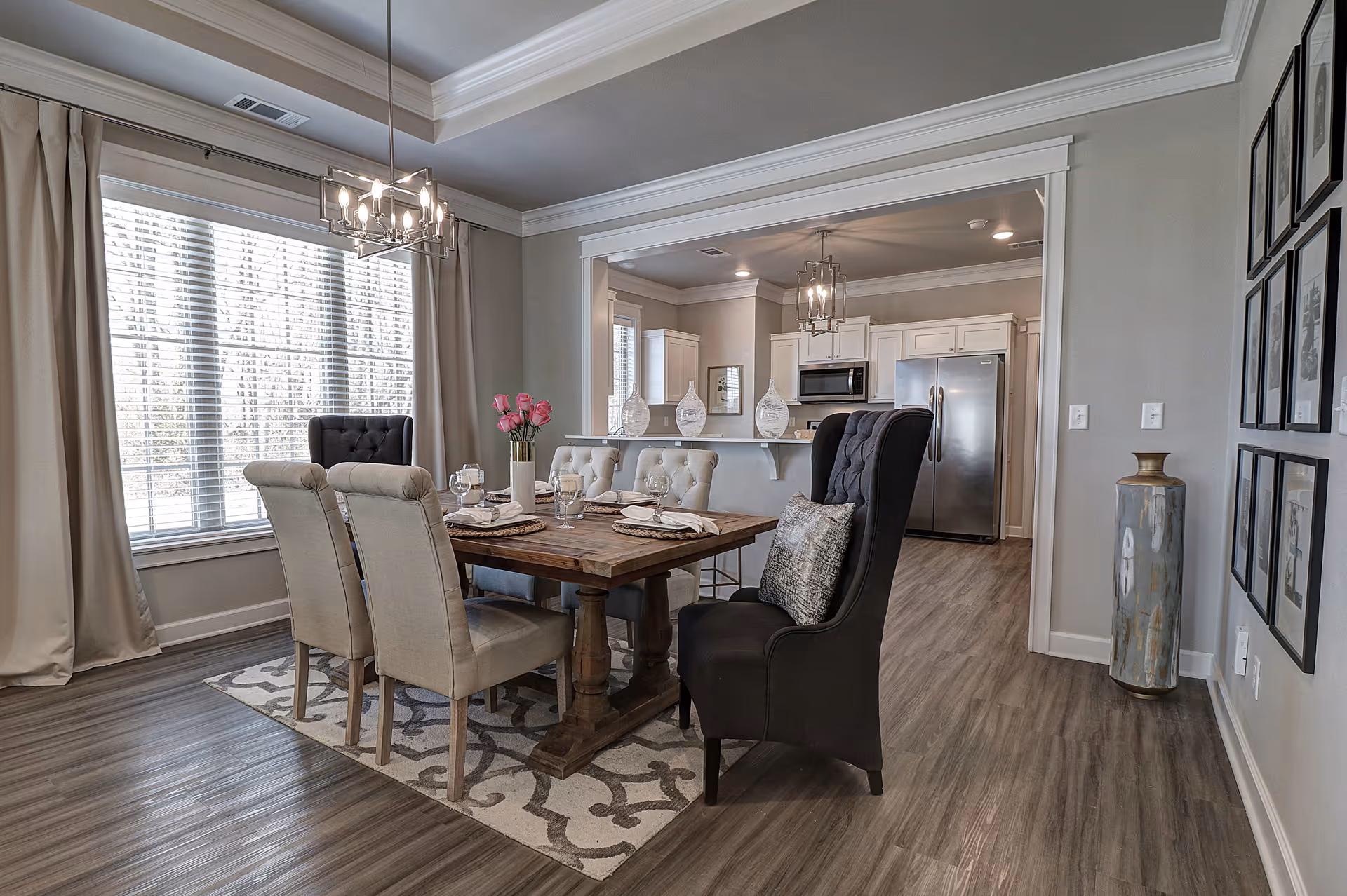 A modern dining room with a wooden dining table set for four, surrounded by upholstered chairs including two high-back black chairs. The room features large windows with beige curtains, a decorative rug under the table, and a chandelier overhead. The dining area opens into a kitchen with white cabinetry, stainless steel appliances, and wood flooring throughout.