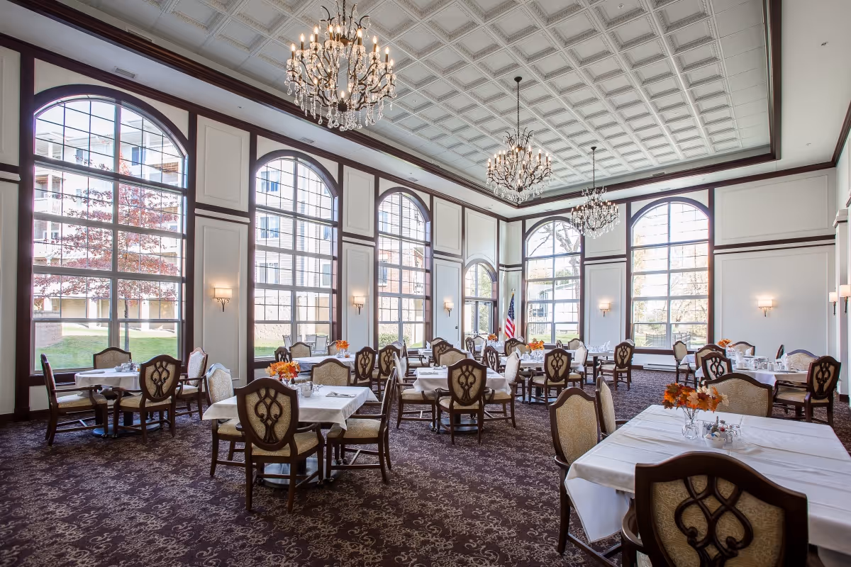 Elegant dining room with large arched windows letting in natural light, multiple tables covered with white tablecloths, ornate chairs, chandeliers hanging from a decorative ceiling, and small floral centerpieces on each table.