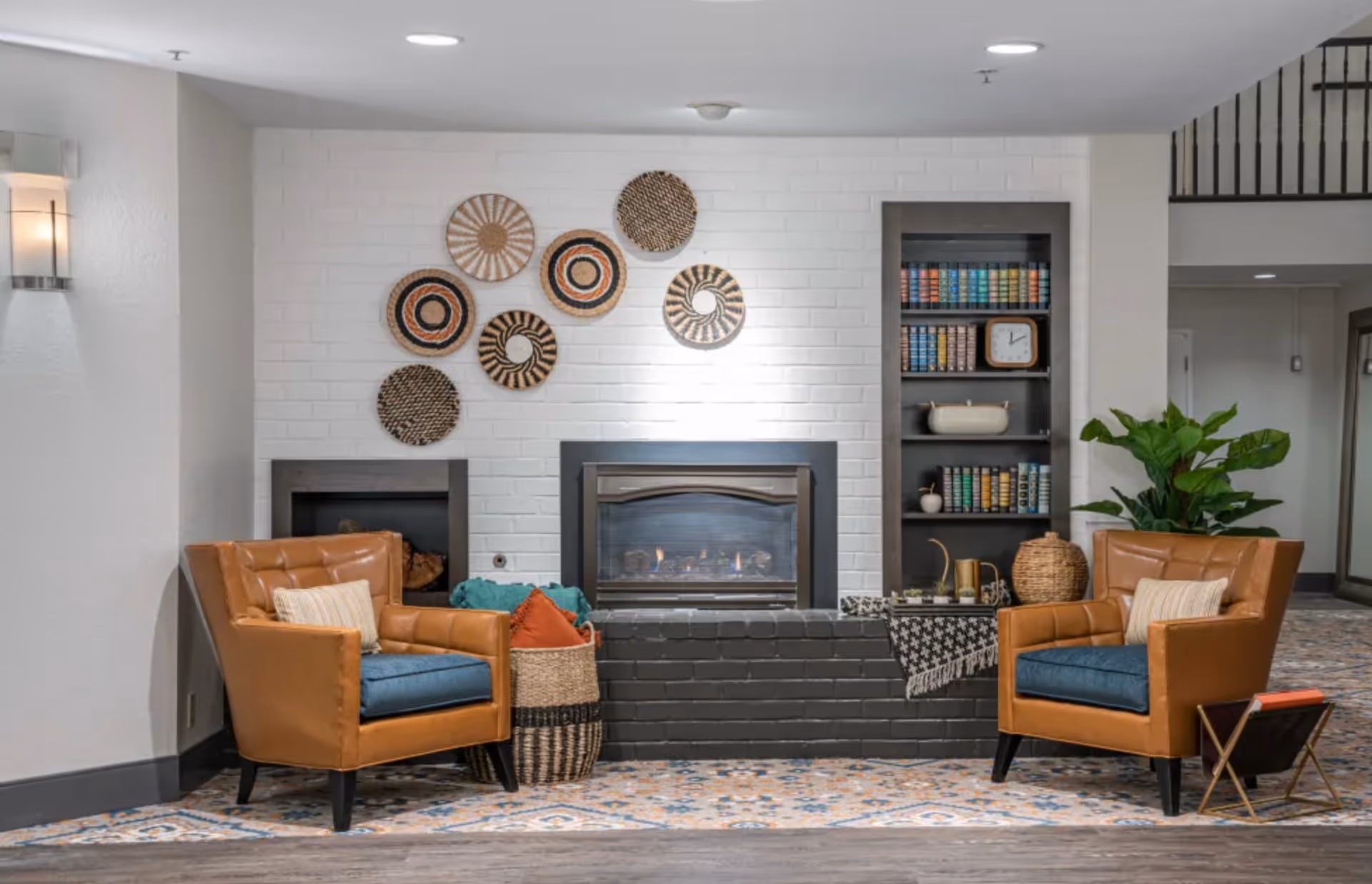 A cozy senior living facility common area with two brown leather armchairs featuring blue cushions and beige pillows, positioned on either side of a black brick fireplace with a fire burning. The white brick wall behind the fireplace is decorated with seven woven circular wall hangings. To the right of the fireplace is a built-in dark wood bookshelf filled with colorful books, a clock, and decorative items. A basket with pillows and a green plant are also visible, along with a patterned carpet and wooden flooring.