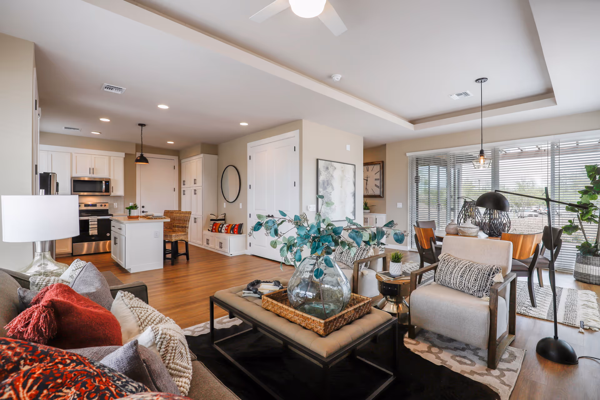 A bright and spacious open-concept living area in a senior living facility featuring a cozy seating area with a sofa, armchair, and coffee table adorned with a large glass vase with greenery. The room includes a modern kitchen with white cabinetry, stainless steel appliances, and a small island with bar stools. Adjacent to the living area is a dining space with a wooden table and chairs near large windows with blinds, allowing natural light to fill the room. The decor is modern and inviting with neutral tones and pops of color from cushions and plants.