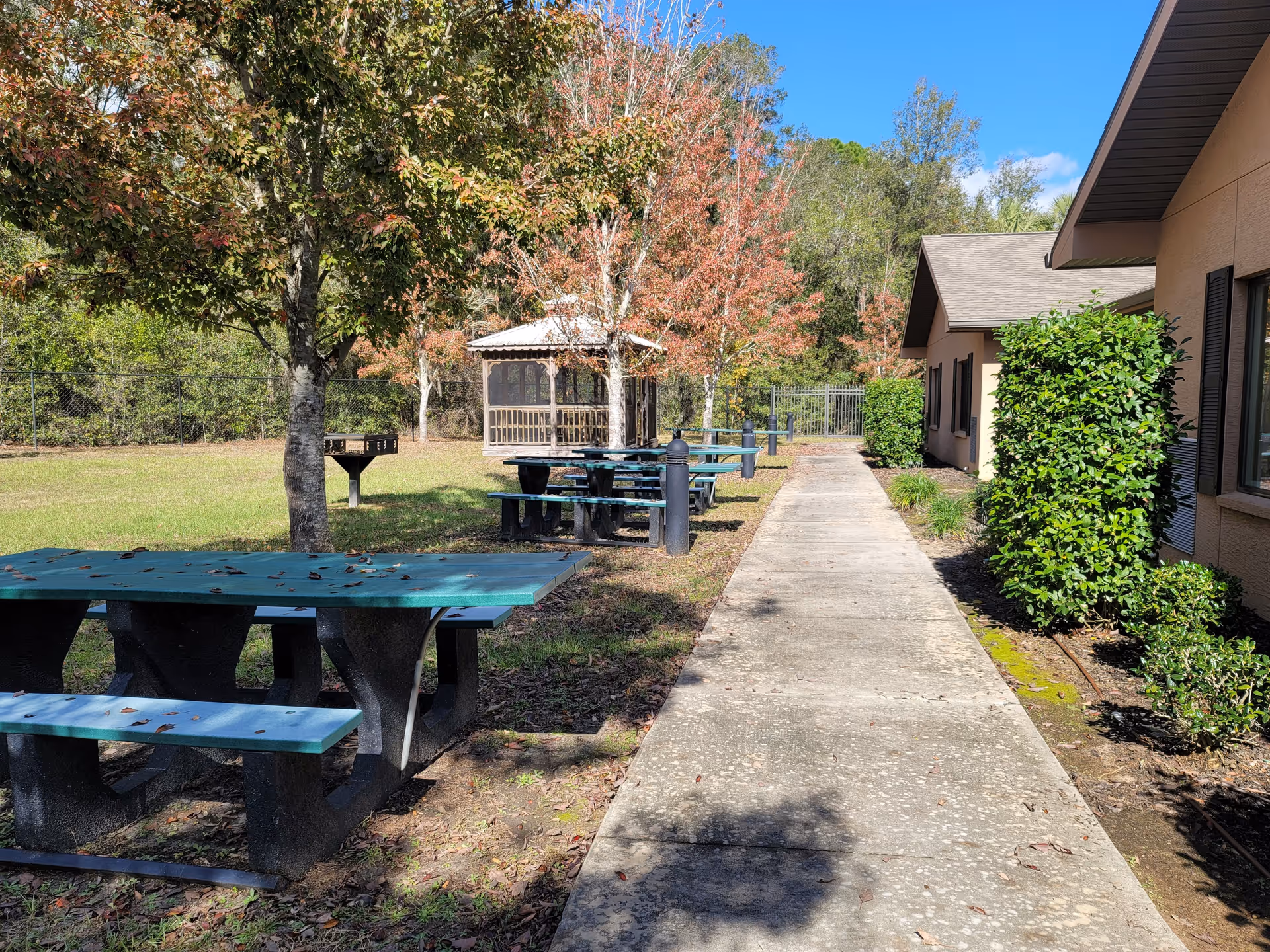 Outdoor area at a senior living facility with a concrete walkway alongside a building, green picnic tables with benches, a barbecue grill, trees with autumn foliage, and a gazebo in the background under a clear blue sky.