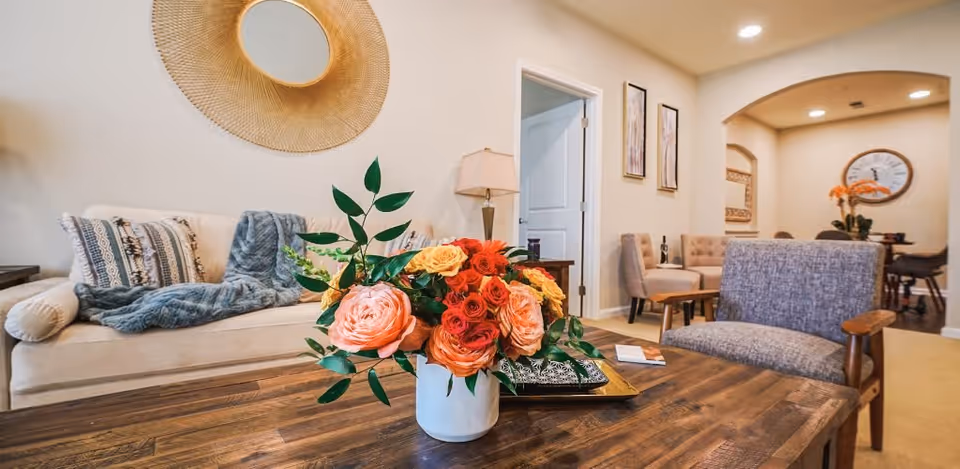 A cozy living room area with a beige sofa adorned with patterned pillows and a blue throw blanket. A wooden coffee table in the foreground holds a white vase filled with vibrant orange, pink, and yellow flowers. To the right, there is a gray upholstered chair with wooden armrests. The background shows a small dining area with chairs, a table, wall art, and a large clock on the wall.