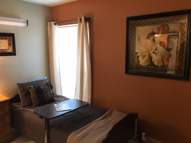 Small private bedroom with a single bed, overbed table, nightstand, window with curtains, and framed artwork on the wall.