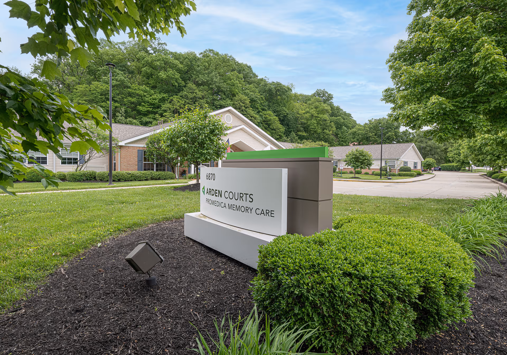 Outdoor view of Arden Courts - ProMedica Memory Care Community sign with surrounding greenery, bushes, trees, and a driveway leading to the facility buildings in the background under a partly cloudy sky.