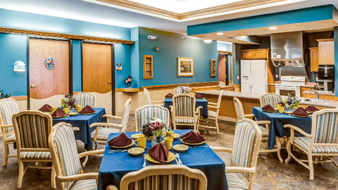 Dining room with several small tables set with blue tablecloths and place settings, upholstered chairs, and an open kitchen area in the background.