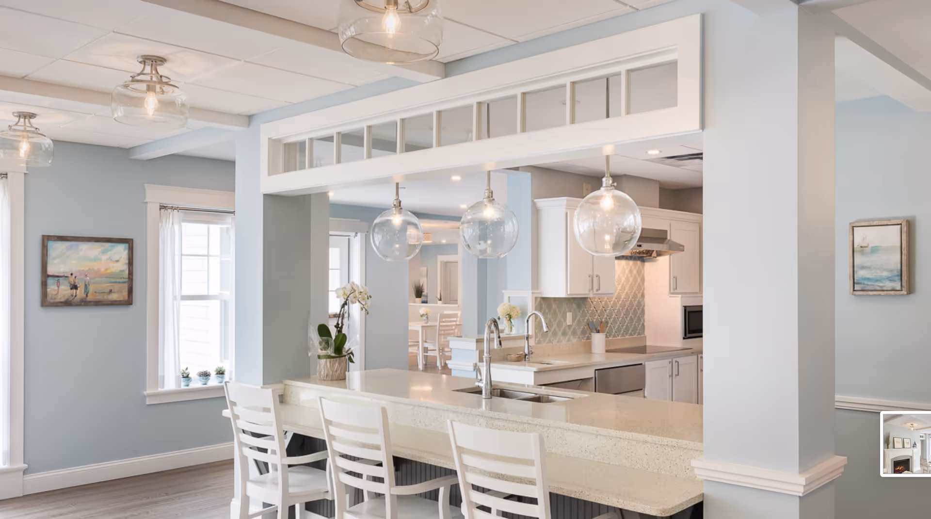 Bright open kitchen with light blue walls, a long island with white chairs and glass pendant lights overlooking an adjacent dining area.