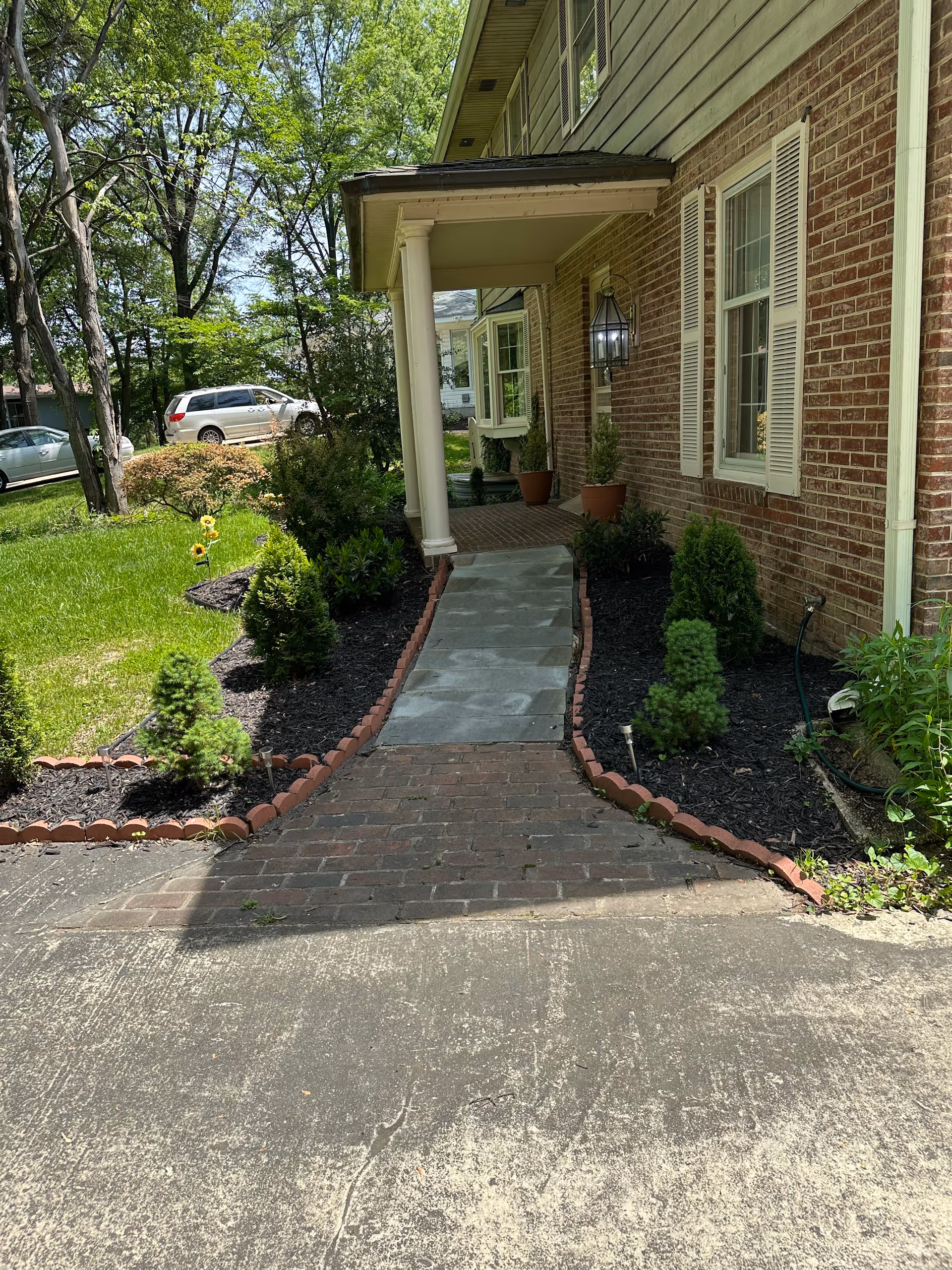 A brick walkway leading to the front porch of a brick house with white shutters and a hanging lantern. The walkway is bordered by small shrubs and mulch beds, with a green lawn and trees visible on the left side. Two cars are parked on the street in the background.