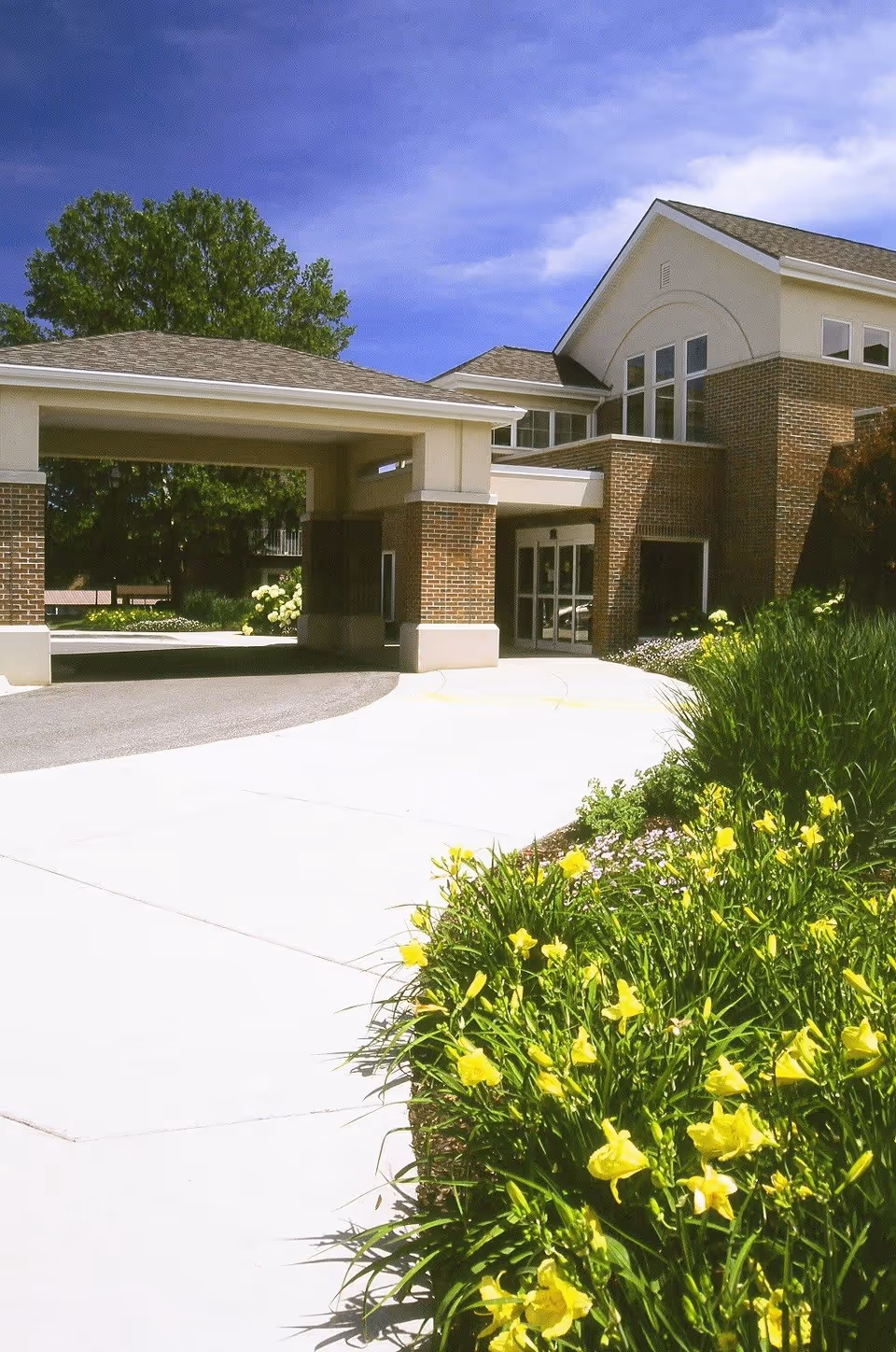 Exterior view of Porter Hills Village building entrance with a covered driveway, brick and beige walls, large windows, and a garden with yellow flowers along the sidewalk under a blue sky.