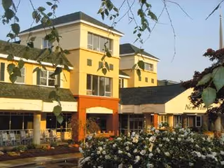 Exterior view of a multi-story senior living facility building with yellow and beige walls, large windows, and a garden with white flowers in the foreground under a clear sky.