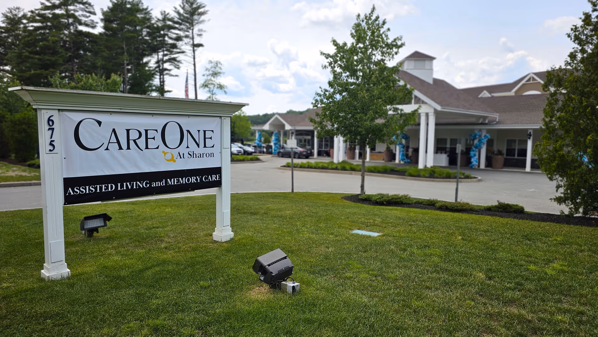 Outdoor view of CareOne at Sharon assisted living and memory care facility with a large sign on a grassy area in front of the building entrance, surrounded by trees and a partly cloudy sky.