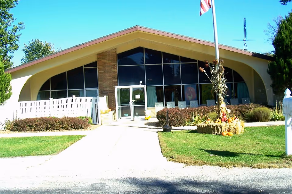 Front entrance of a mid-century single-story building with a steep gabled roof, large glass windows, a flagpole, and fall decorations on the lawn.