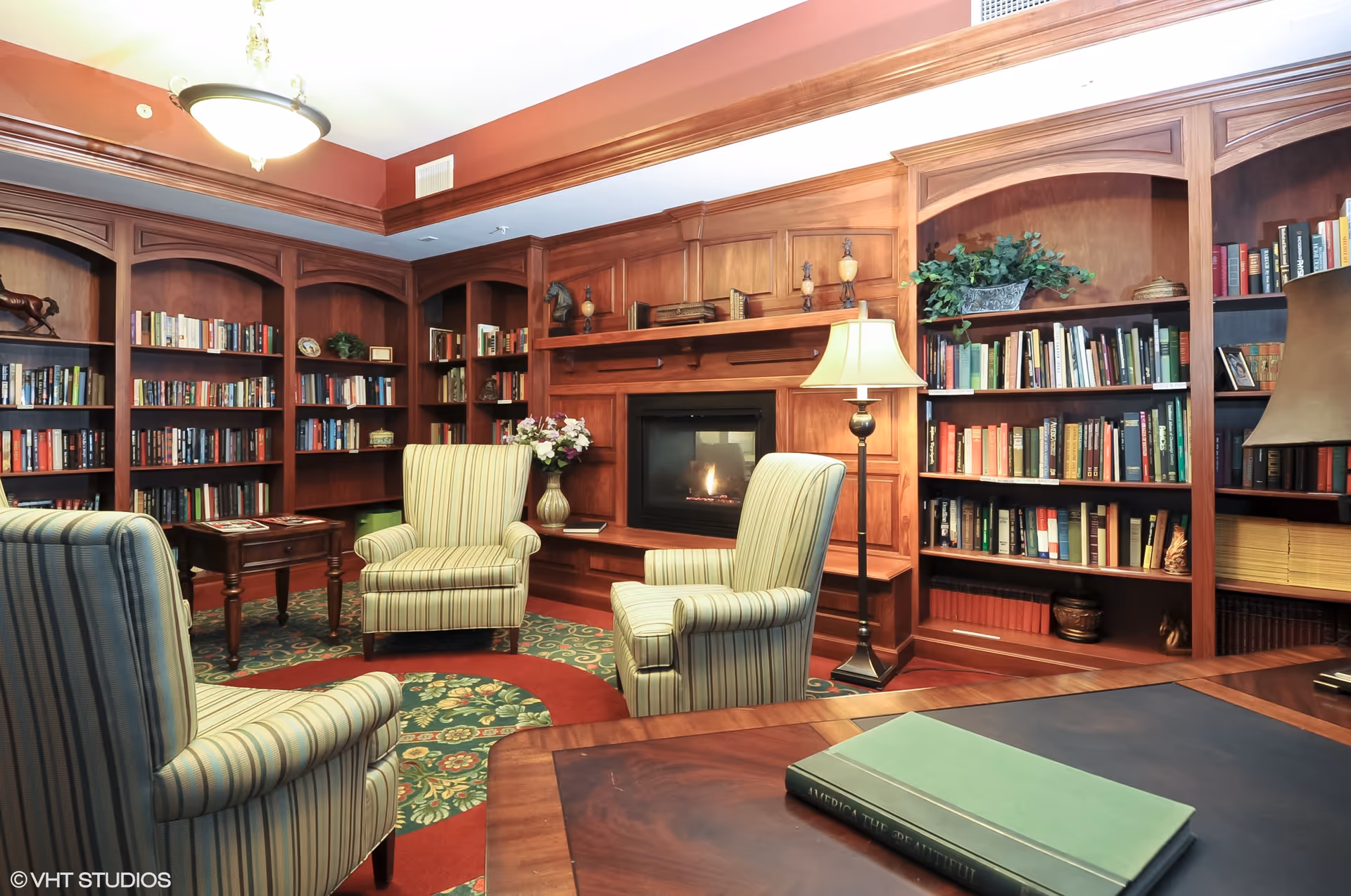 A cozy library room with wooden bookshelves filled with books and decorative items. There are three striped upholstered armchairs arranged around a fireplace with a lit fire. A floor lamp stands beside one of the chairs, and a wooden table with a green book is in the foreground. The room has warm lighting and a patterned carpet.