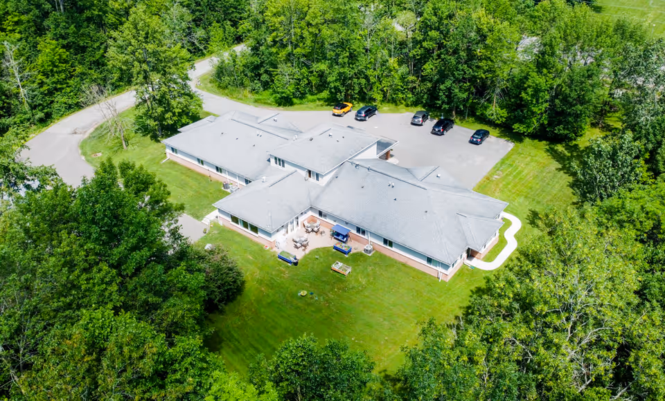 Aerial view of a single-story senior living facility surrounded by dense green trees and grass. The building has a gray roof and a small parking lot with several cars parked. There is a curved walkway leading to the entrance and outdoor seating areas on the lawn.