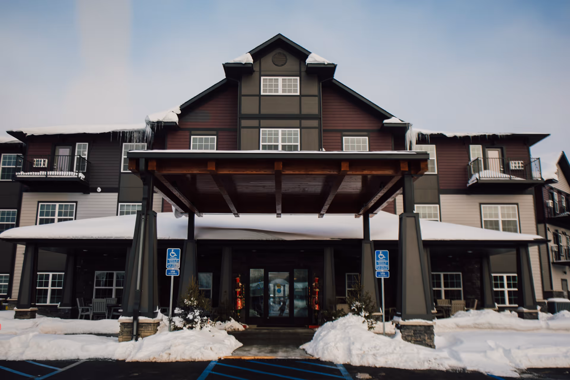 Front exterior view of a multi-story senior living facility building with a covered entrance, snow on the ground and roof, and two handicap parking signs near the entrance.