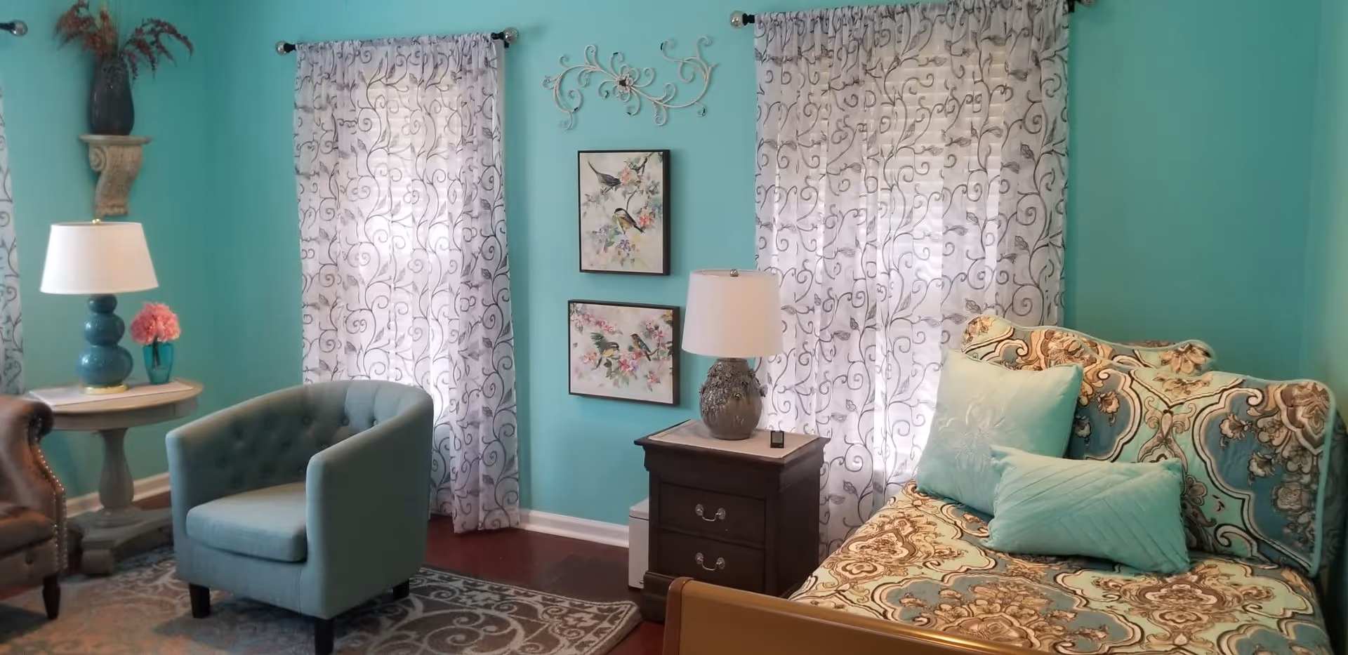 A cozy living room with turquoise walls, two windows with sheer patterned curtains, a patterned sofa with turquoise pillows, a turquoise armchair, a small wooden side table with a lamp, and wall art featuring birds and floral designs.