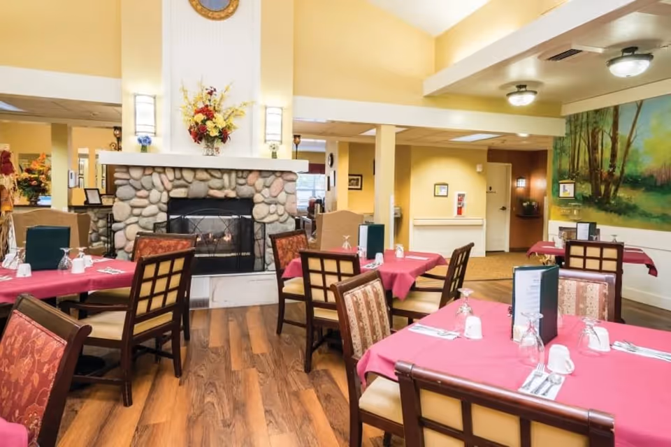Dining room with several tables covered in red tablecloths, set with glasses, cups, and silverware. The room features a stone fireplace with a floral arrangement on the mantel, wooden chairs with patterned cushions, and a large mural of a forest scene on one wall.