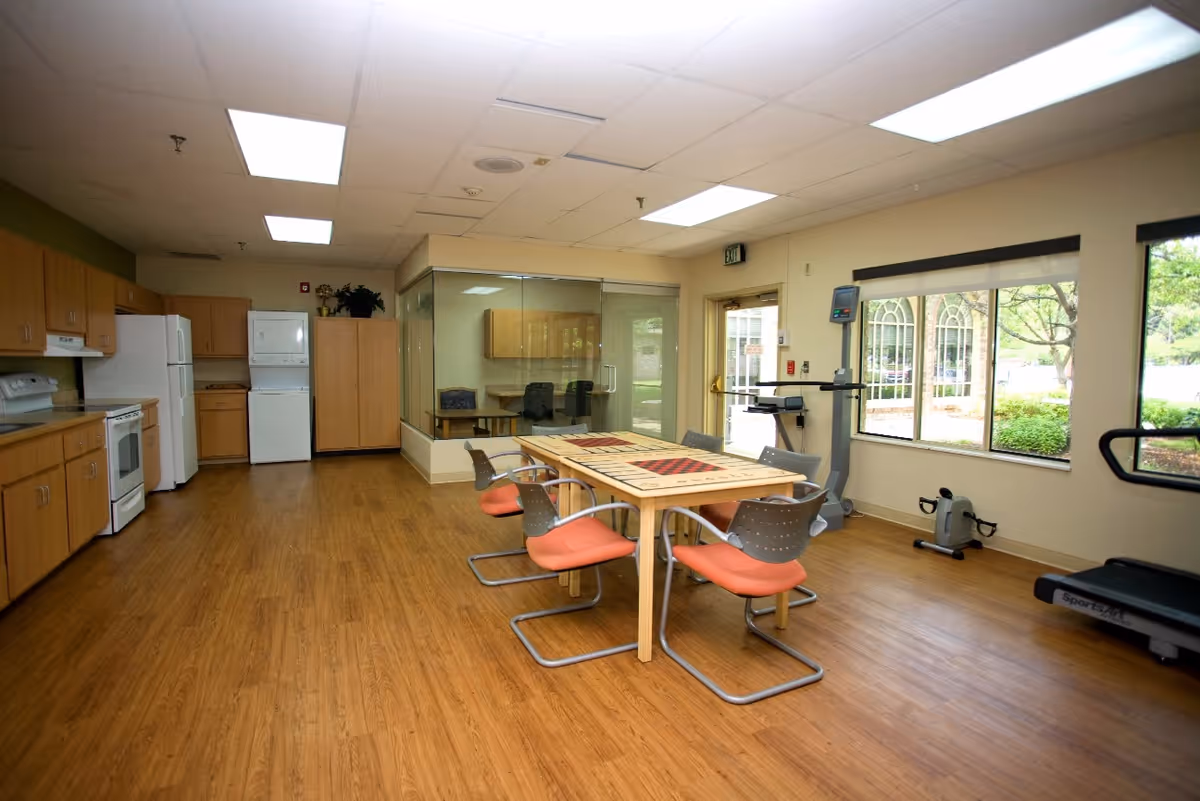 Bright communal activity room with a central table and chairs, a kitchenette along the left wall, and exercise equipment by the windows.