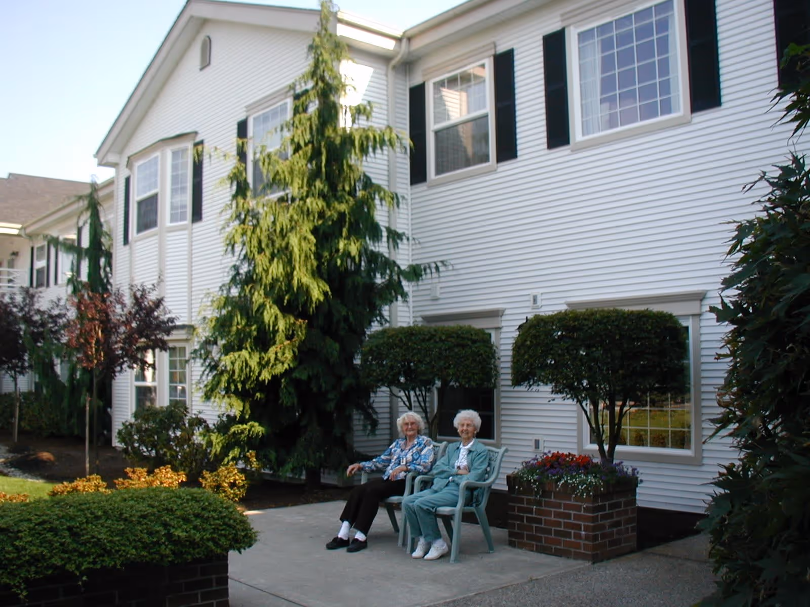 Two elderly women sitting on chairs outside a white residential building with large windows, surrounded by well-maintained bushes, trees, and flower beds.