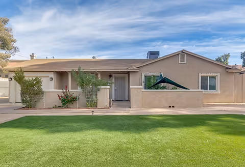 Single-story desert-themed senior living facility, beige exterior, green lawn, and shaded patio area.