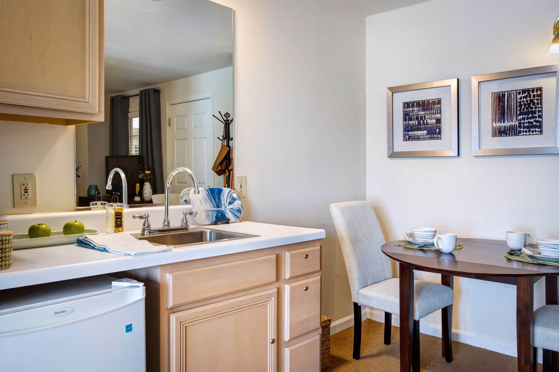 Small kitchenette with a sink and countertop beside a round dining table set for two and framed artwork on the wall.