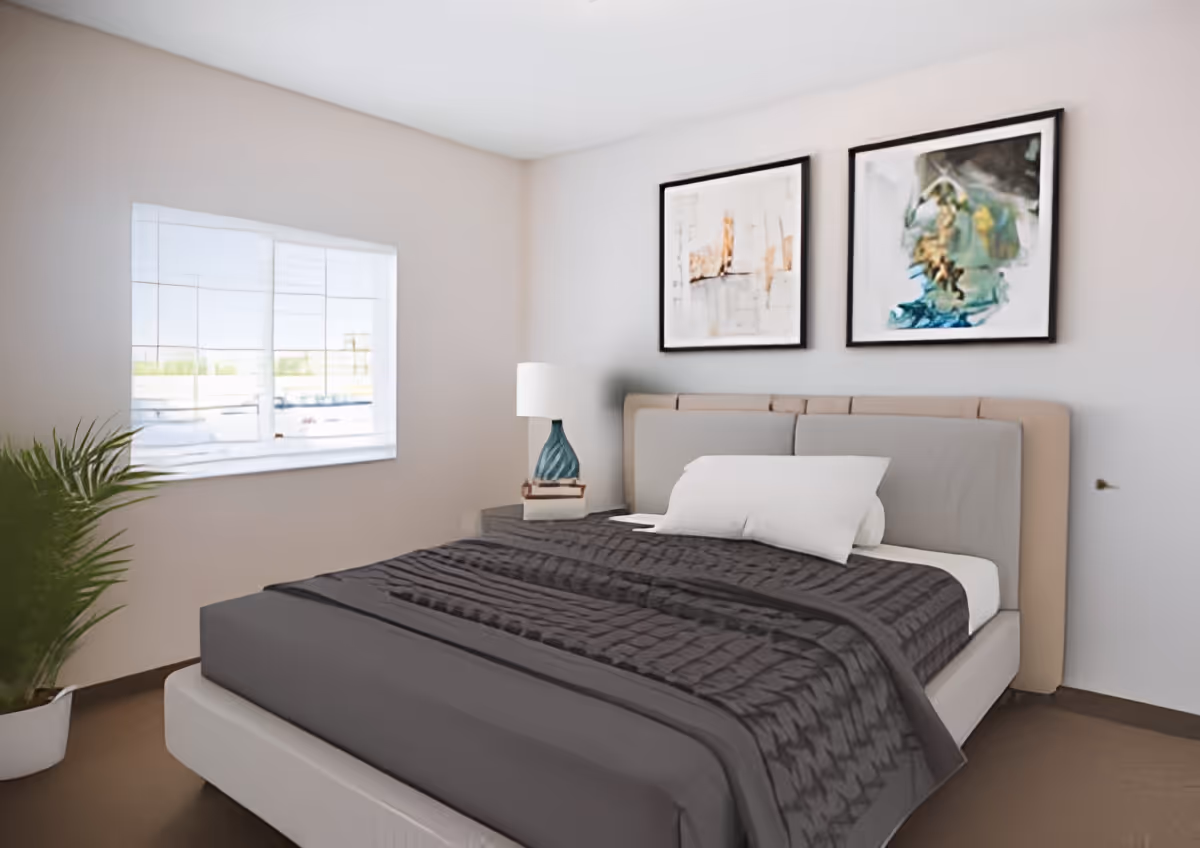 A modern, softly lit bedroom with a double bed made with a dark gray quilt, a bedside table and lamp, framed artwork above the headboard, and a potted plant by the window.