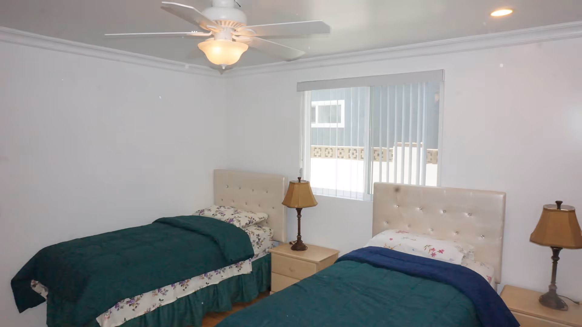 A bedroom with two single beds, each with a tufted beige headboard, floral sheets, and dark green blankets. Between the beds is a small wooden nightstand with a lamp. Another lamp is on a small table next to the right bed. A window with vertical blinds is on the wall behind the beds, and a ceiling fan with a light fixture is mounted on the white ceiling.