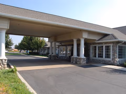 Entrance area of a senior living facility with a covered driveway supported by white columns and stone bases, adjacent to a building with large windows and stone accents under a clear sky.
