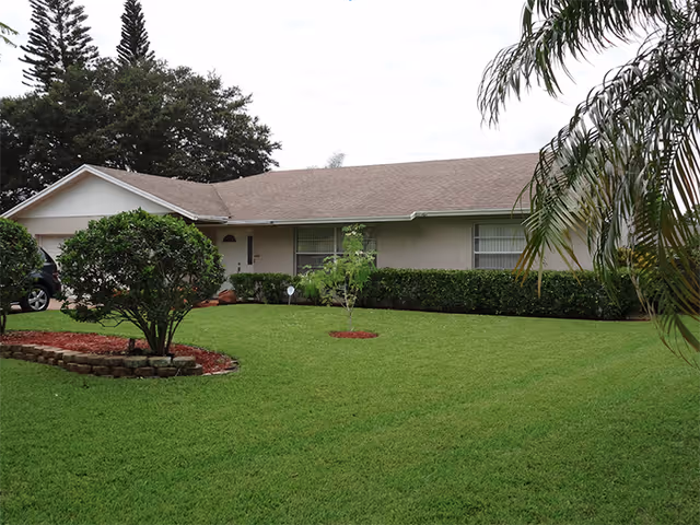 Single-story beige ranch-style house with a manicured green lawn, shrubs, and a garage in front.