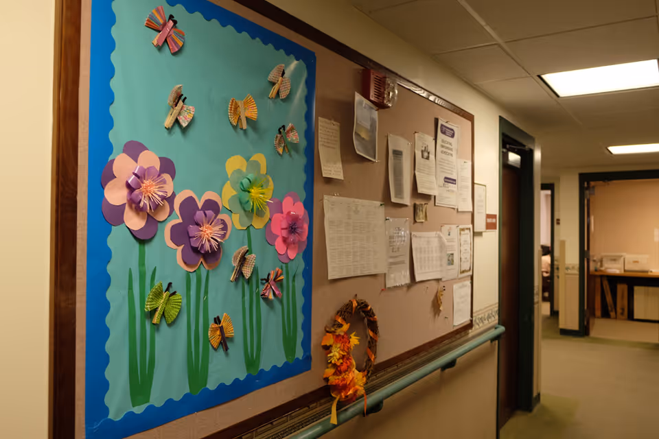 A nursing home corridor with a bulletin board decorated with colorful paper flowers and posted notices.