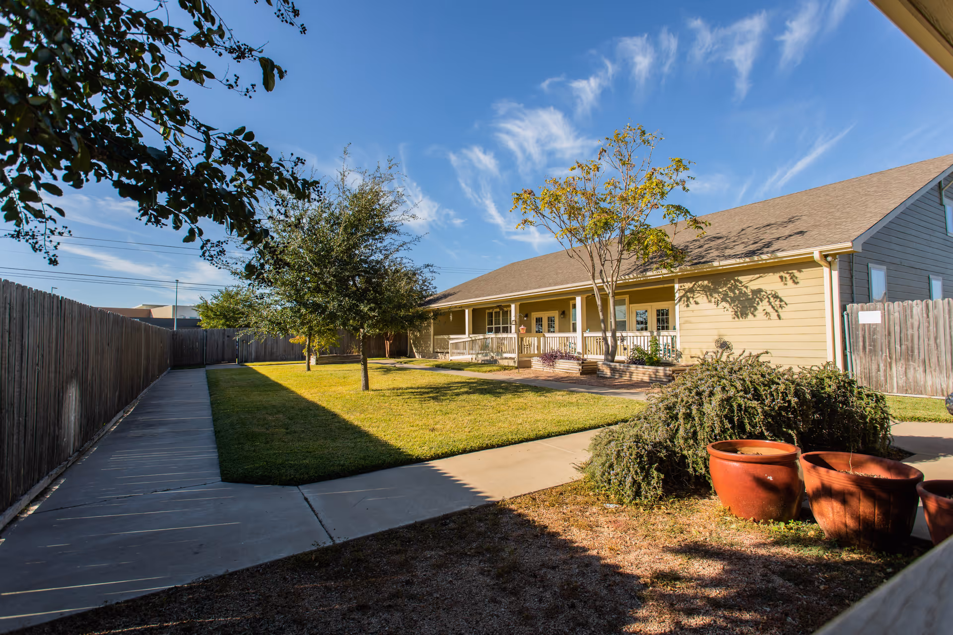 Outdoor view of a single-story building with a beige exterior and a sloped roof, surrounded by a fenced yard with a concrete walkway, green grass, trees, and large terracotta pots in the foreground under a blue sky with wispy clouds.