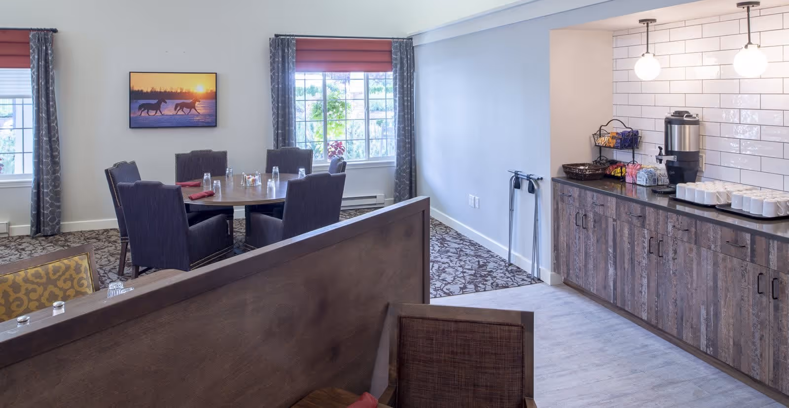 A dining area in Kipling Meadows Senior Living featuring a round table with six chairs, set with glasses and napkins. The room has two windows with patterned curtains, a wall-mounted picture of horses at sunset, and a side counter with a coffee dispenser, cups, and snacks. The flooring is a combination of carpet and light wood, and the walls are painted in soft neutral tones.