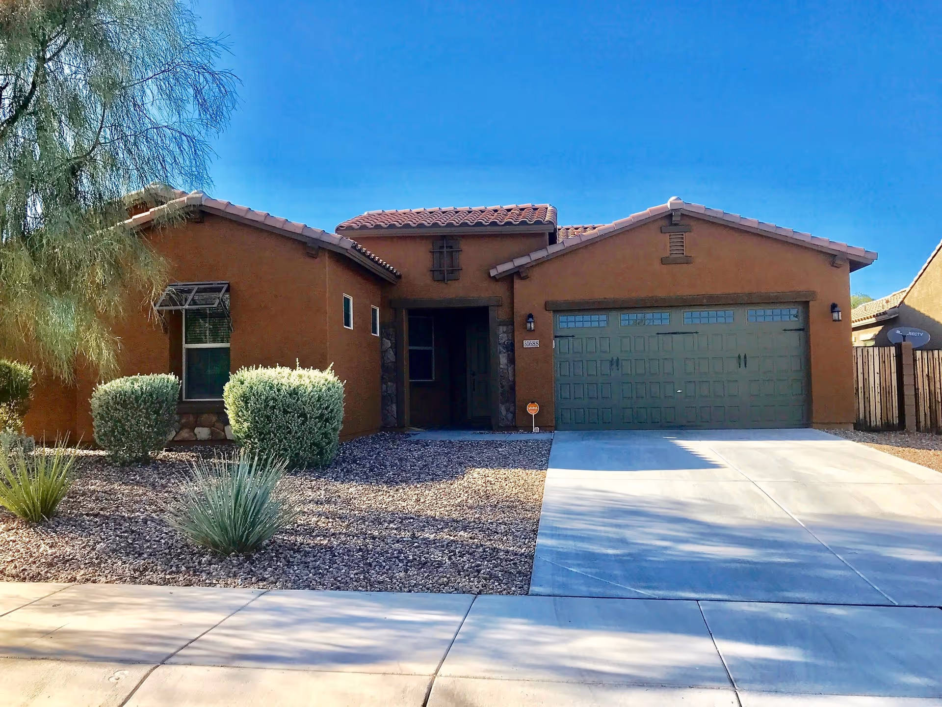 Front exterior view of a single-story house with a brown stucco finish, a tiled roof, a two-car garage with green doors, and a driveway. The front yard features desert landscaping with gravel, bushes, and a tree under a clear blue sky.
