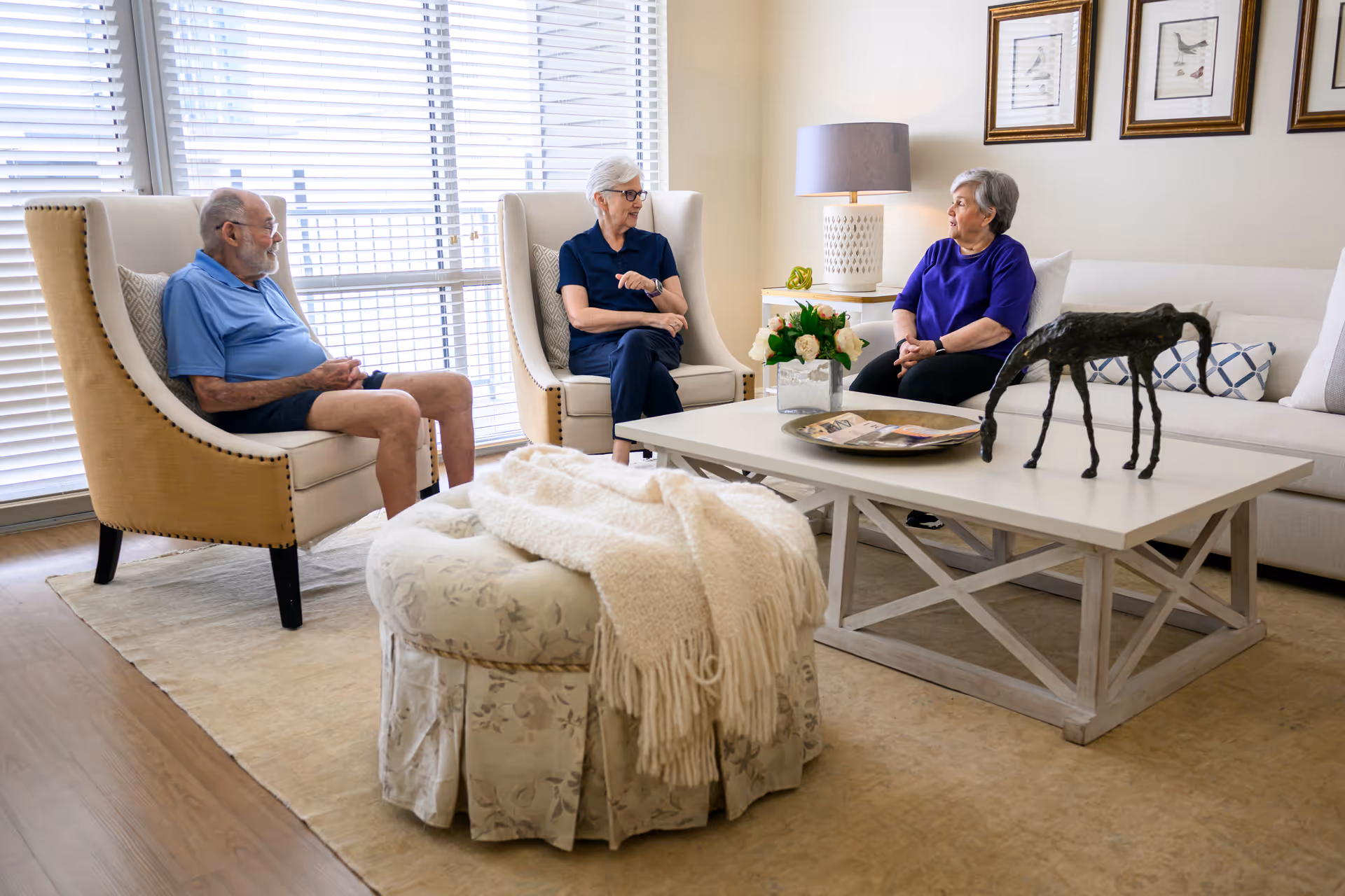 Three elderly people sitting and conversing in a bright living room with large windows covered by blinds. The room features beige armchairs, a white coffee table with decorative items, a floral ottoman with a cream-colored throw blanket, and framed artwork on the wall.