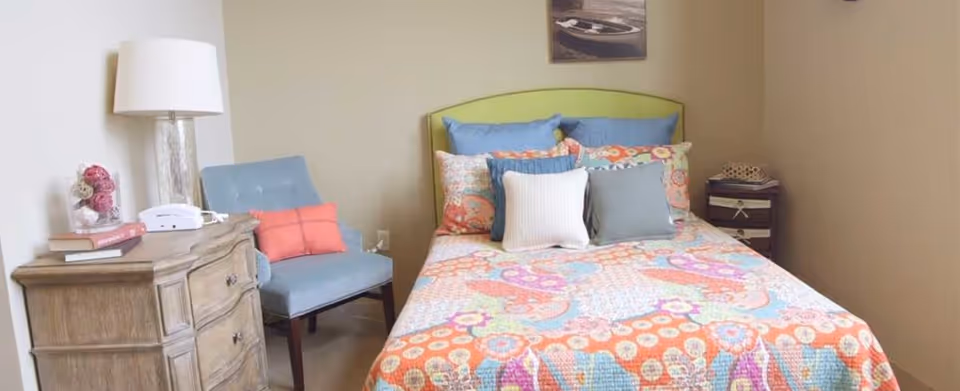 A cozy bedroom featuring a bed with a colorful patterned quilt and multiple pillows in shades of blue, white, and orange. To the left of the bed is a wooden nightstand with a white lamp, books, and a decorative item. A light blue upholstered chair with an orange cushion is positioned next to the nightstand. The walls are painted beige, and there is a small piece of artwork hanging above the bed.