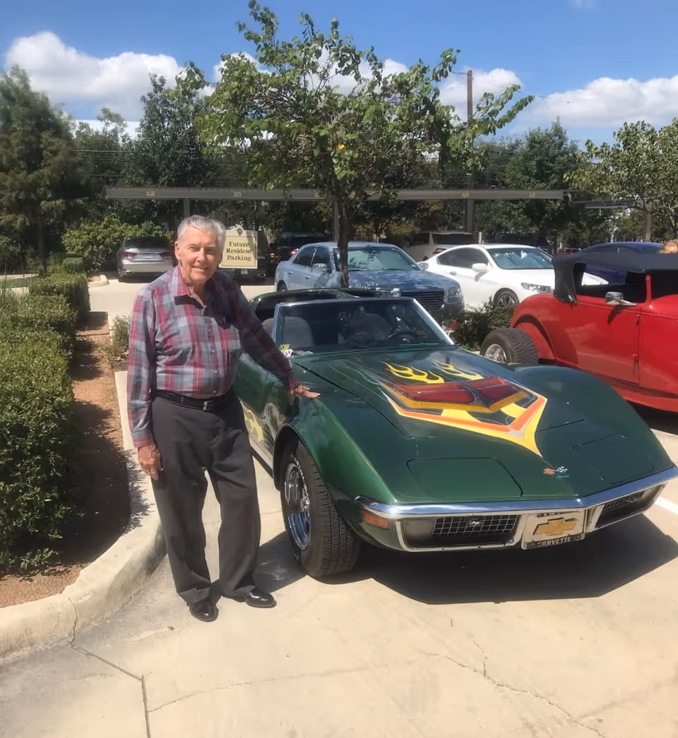 An elderly man wearing a plaid shirt and dark pants stands next to a green vintage Chevrolet Corvette with a colorful flame design on the hood in a parking lot. Several other cars and trees are visible in the background under a partly cloudy sky.