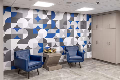 Two blue armchairs and a small table sit against a geometric patterned accent wall in a senior living common area.